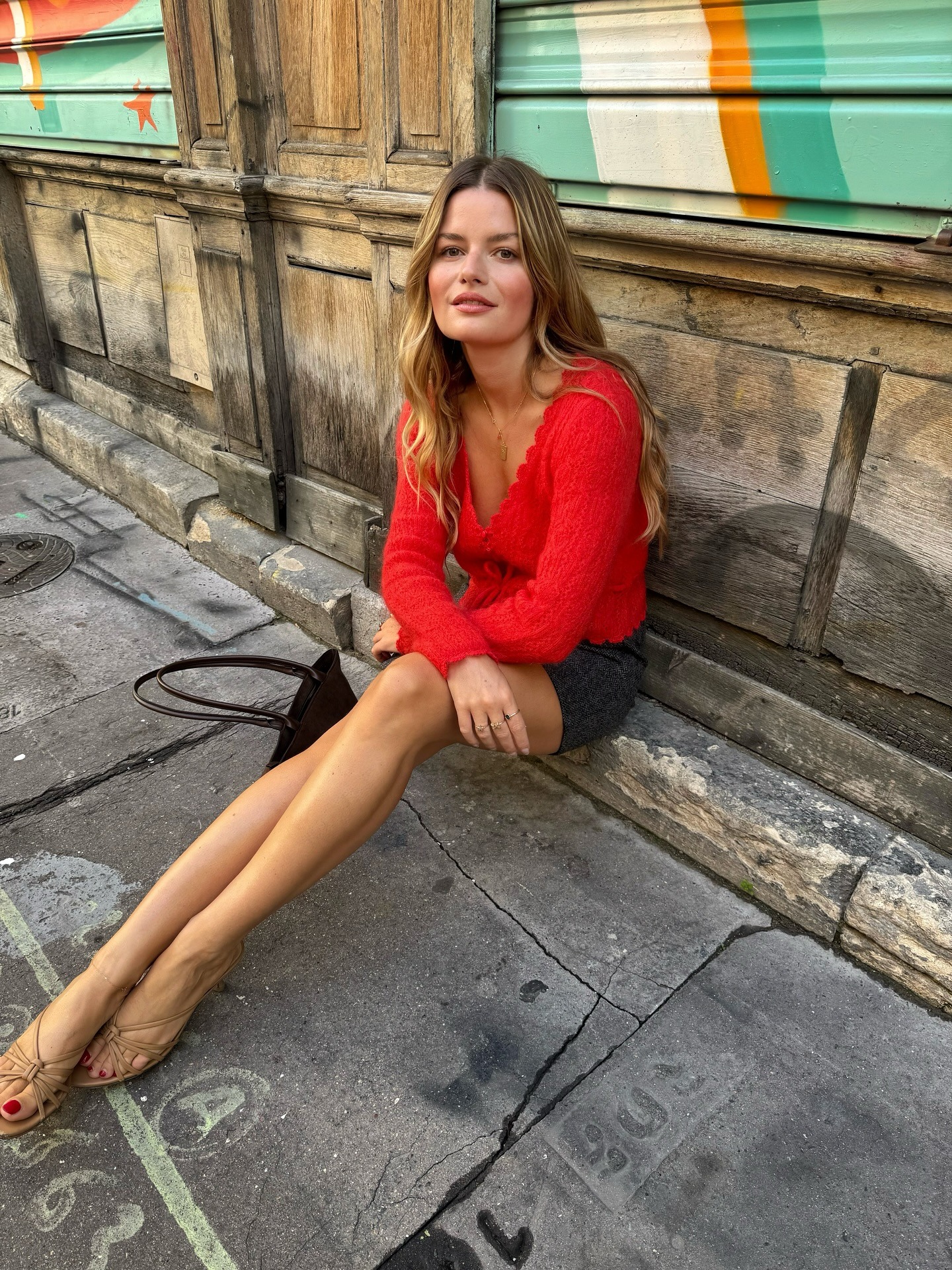 A woman in Paris sitting on a side step wearing a red cardigan with warm, honey blonde hair