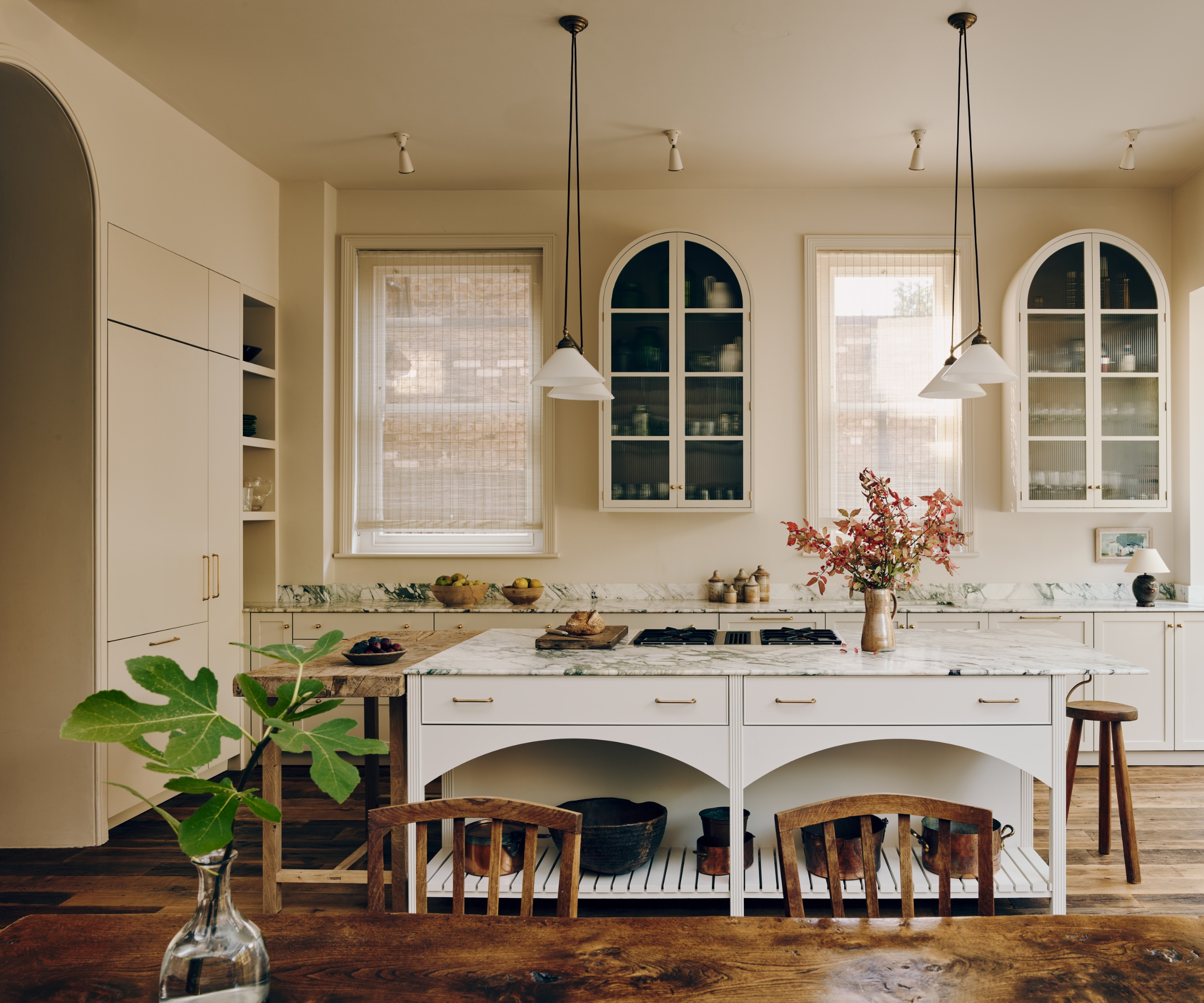 A warm neutral kitchen with curved wall cabinets with glazed fronts, a rectangular white island with curved details below the drawers and topped with marble countertops