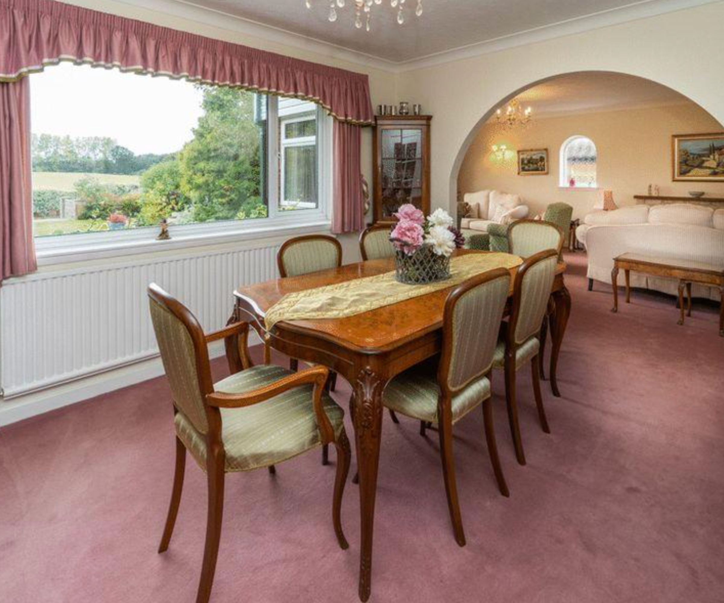 Laura's former dining room with a long wooden table, pale red carpet, and frilly curtains on a window overlooking fields and the garden