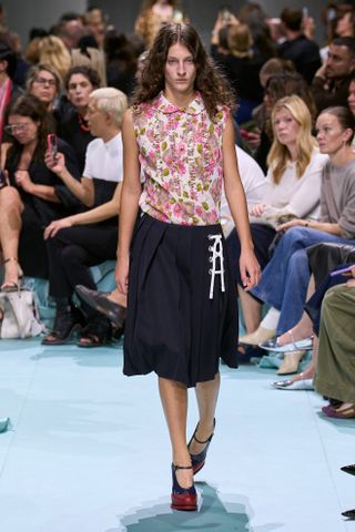 Model wears a pleated skirt on the Prada S/S 25 runway with a floral tank top, and platform Mary Jane shoes.