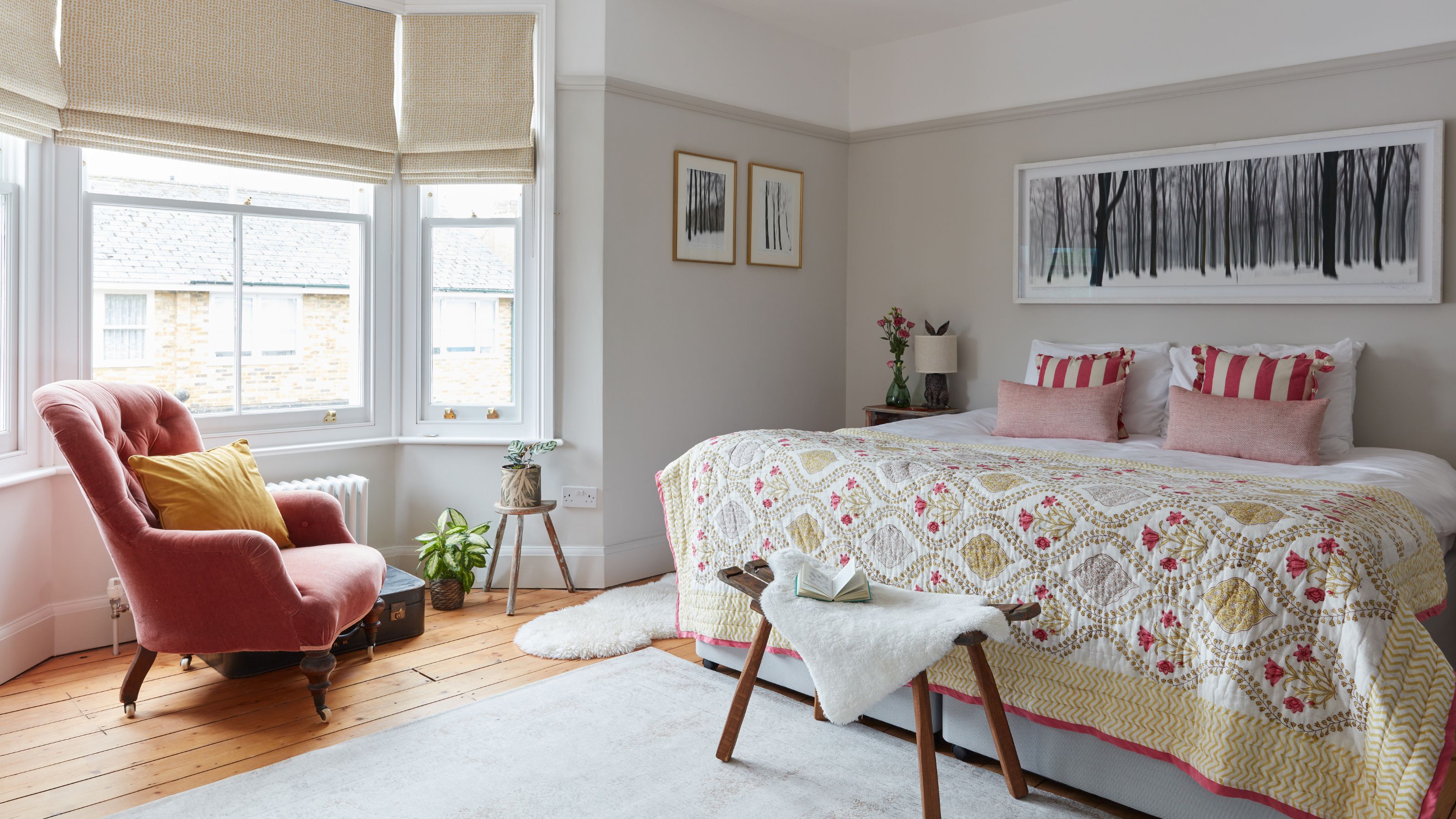 Cream painted bedroom with a bay window, artwork above the bed, and a pink accent chair in front of the bed