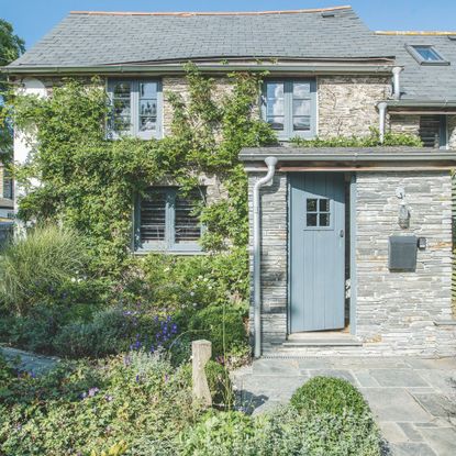 Grey shingle clad house with a blue front door and a wild front garden with plants growing up the front of the house