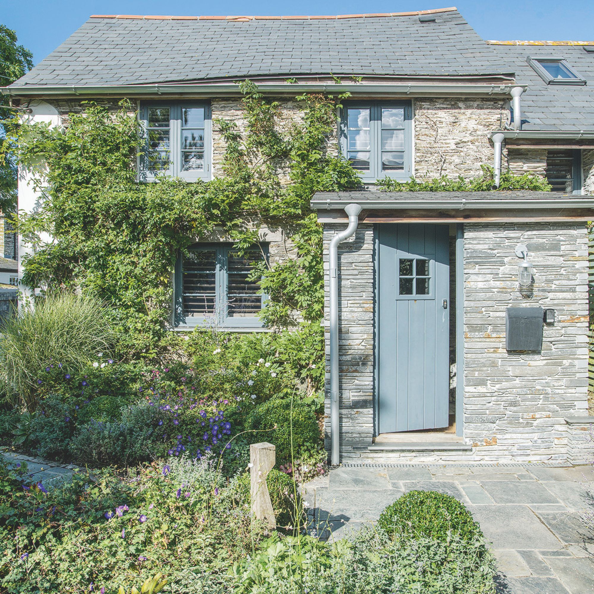 Grey shingle clad house with a blue front door and a wild front garden with plants growing up the front of the house