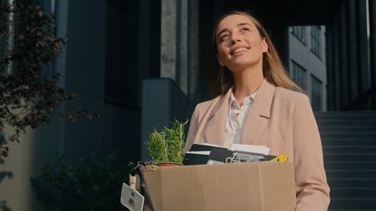 An optimistic-looking woman leaves an office building with a box full of personal items.