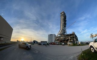 A sunrise, on left, illuminates a blue sky at NASA's Kennedy Space Center. In the foreground, right, is the mobile launcher 1 towering above the ground. The Vehicle Assembly Building is seen in back.