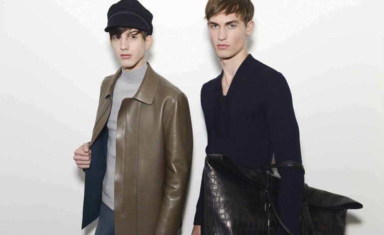 Two male models wearing looks from Gucci&#039;s collection. One model is wearing a dark hat, grey turtle neck jumper and dark sage coloured jacket. The other model is wearing a blue V-neck style jumper and is holding an oversized black bag