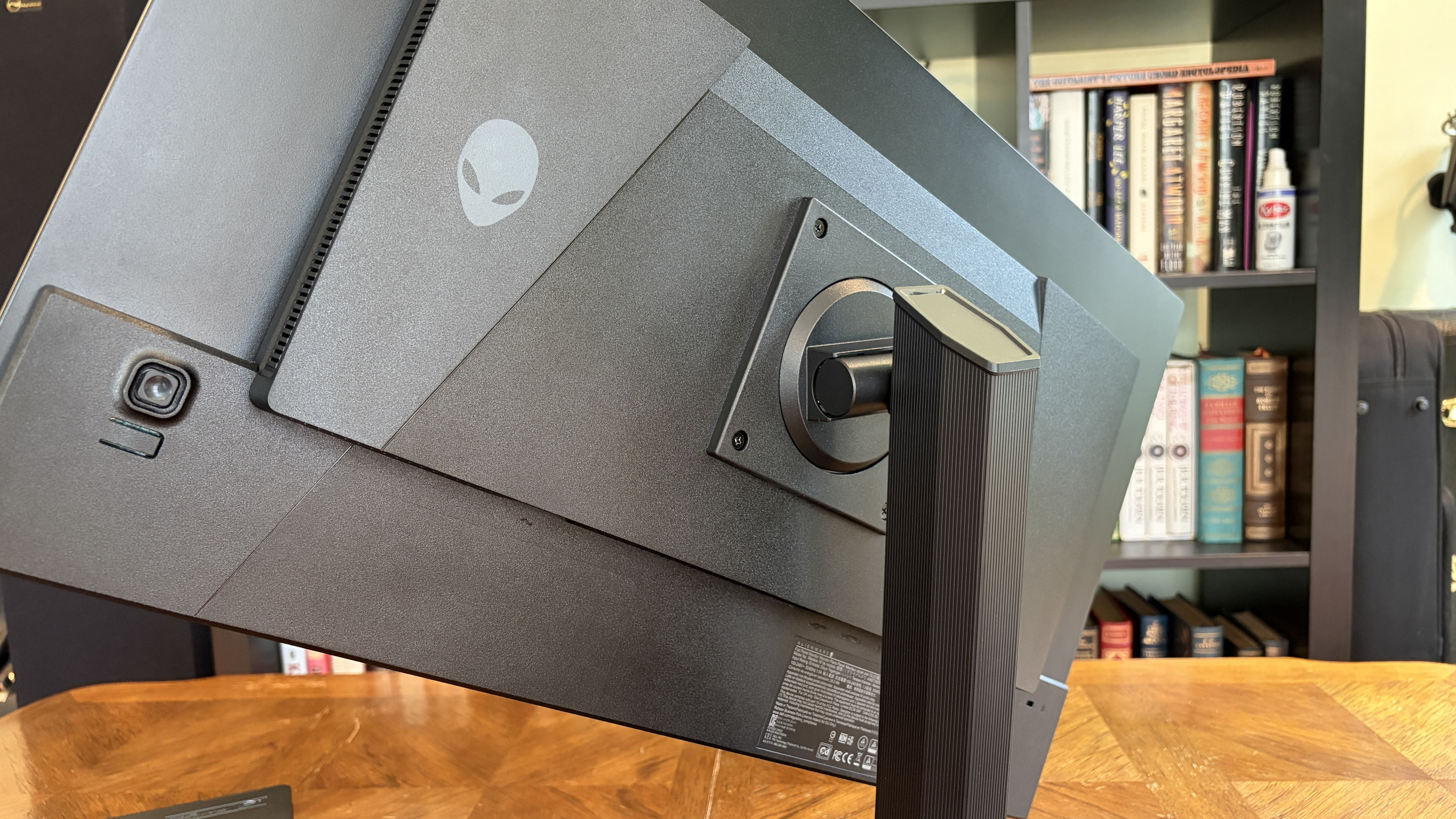 A black computer monitor with a sleek, adjustable stand on a wooden desk. A bookshelf with various books is in the background, adding a cozy atmosphere.