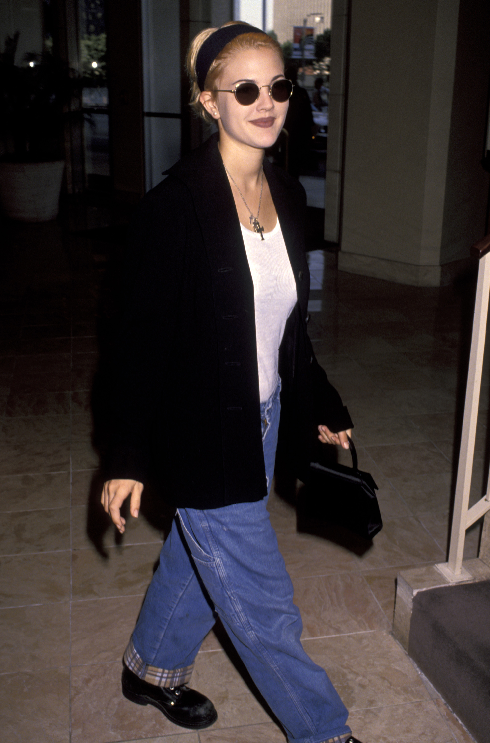 Drew Barrymore wears a 90s autumn jean outfit. Pictured in front of the Century Plaza Hotel in 1992 she wears a oversized black blazer, a white tank, blue jeans and black leather loafers. She finishes the look with oval sunnies and a black micro bag. This image was taken from Getty images recently.