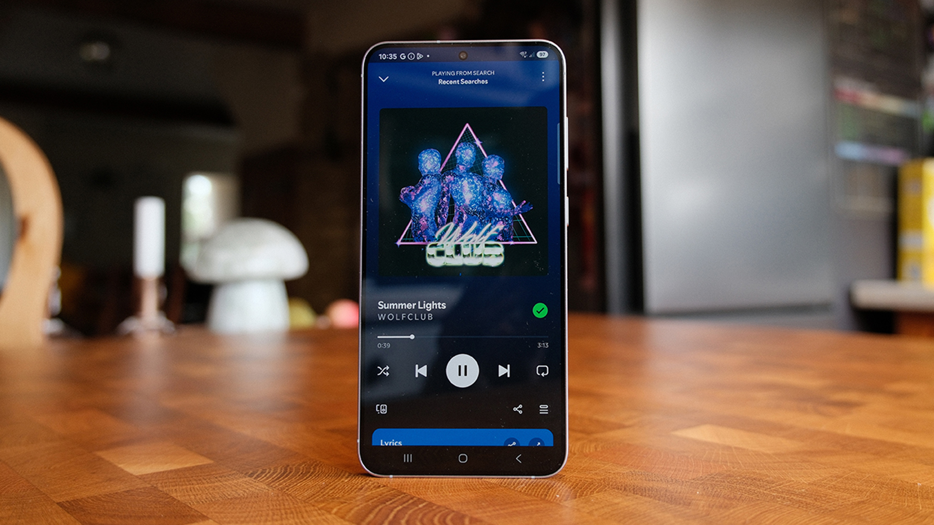 Samsung Galaxy S25 Plus smartphone stood up on wooden table showing music player on screen