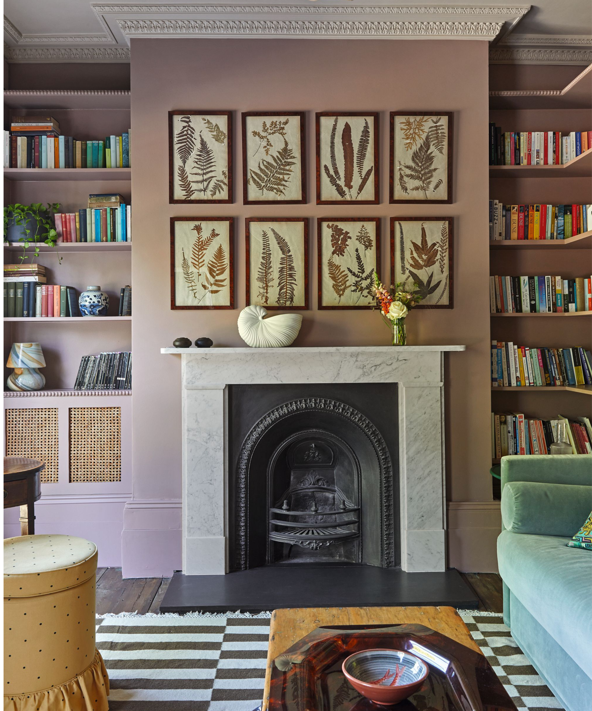 a living room with a black and white striped rug, a green couch, a wooden coffee table, a fireplace with a marble mantel, pink painted walls, artwork hanging above the fireplace, and shelves holding lots of books