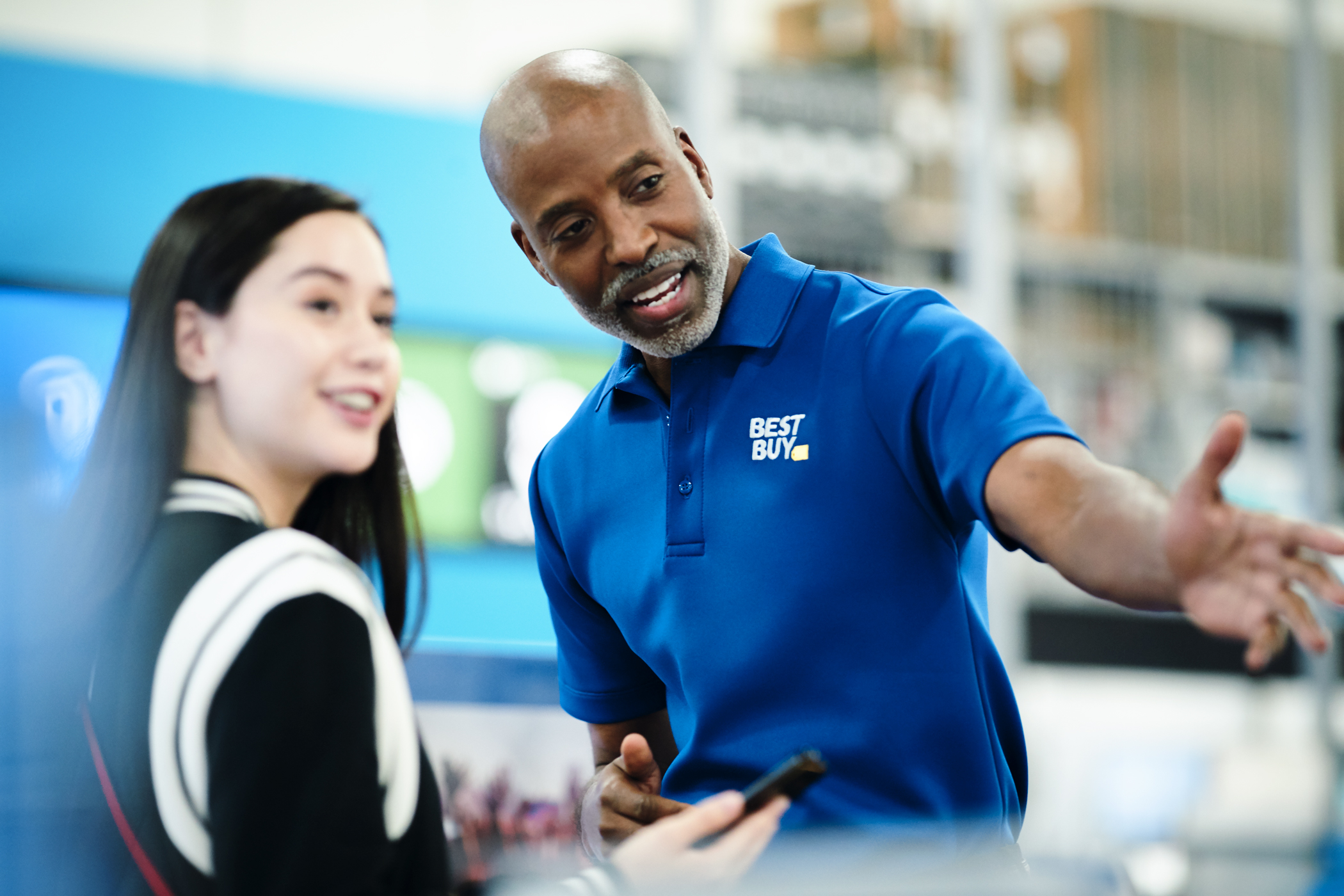 A smiling Best Buy employee in a blue polo shirt gestures with his hand while talking to a customer in a store aisle.