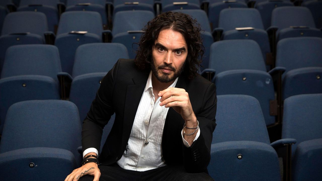 Comedian Russell Brand poses in a suit seated in a theatre seating area