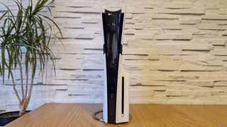 The PS5 Pro console on a wooden desk in front of a white brick background and next to a potted plant