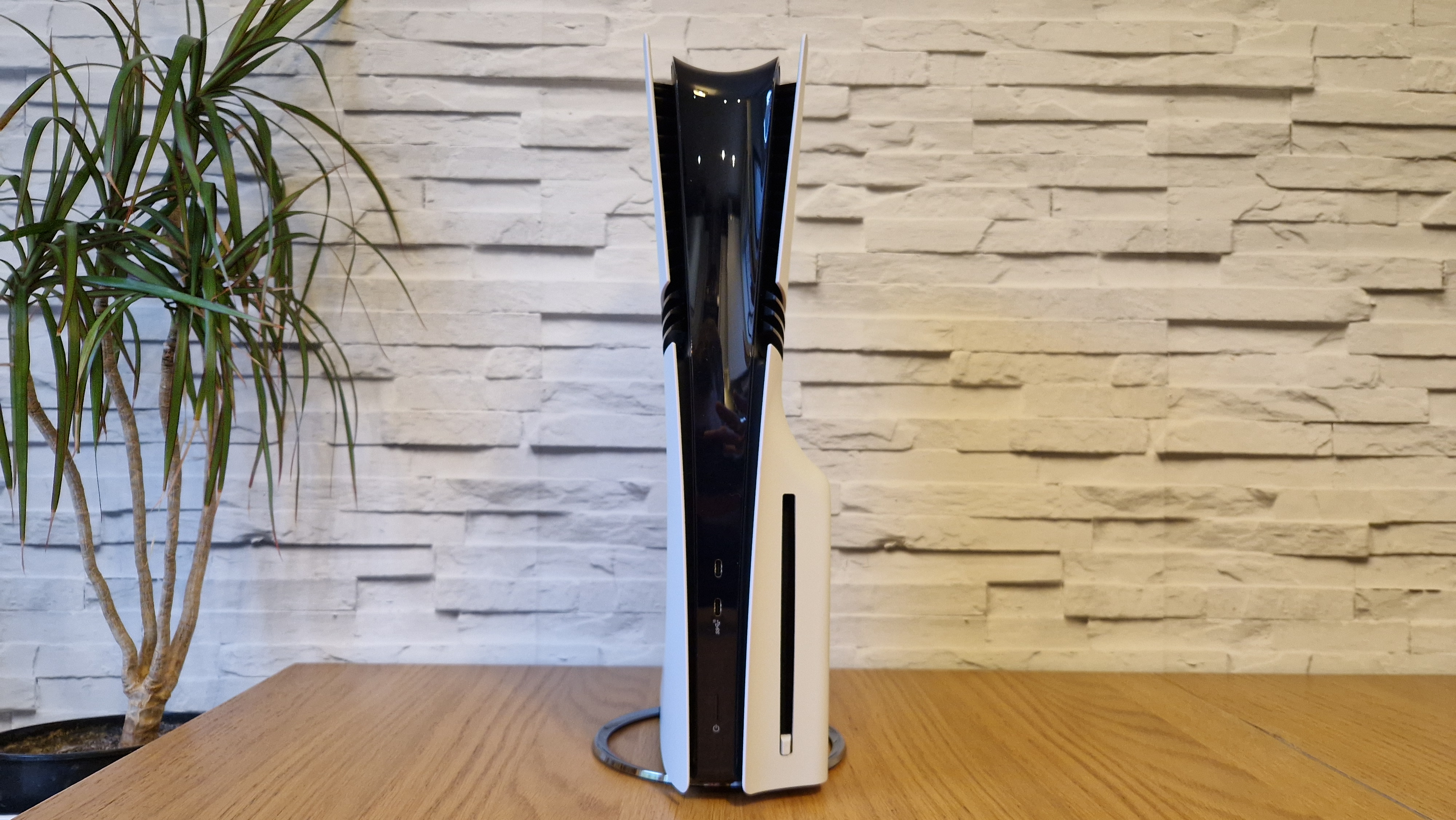 The PS5 Pro console on a wooden desk in front of a white brick background and next to a potted plant