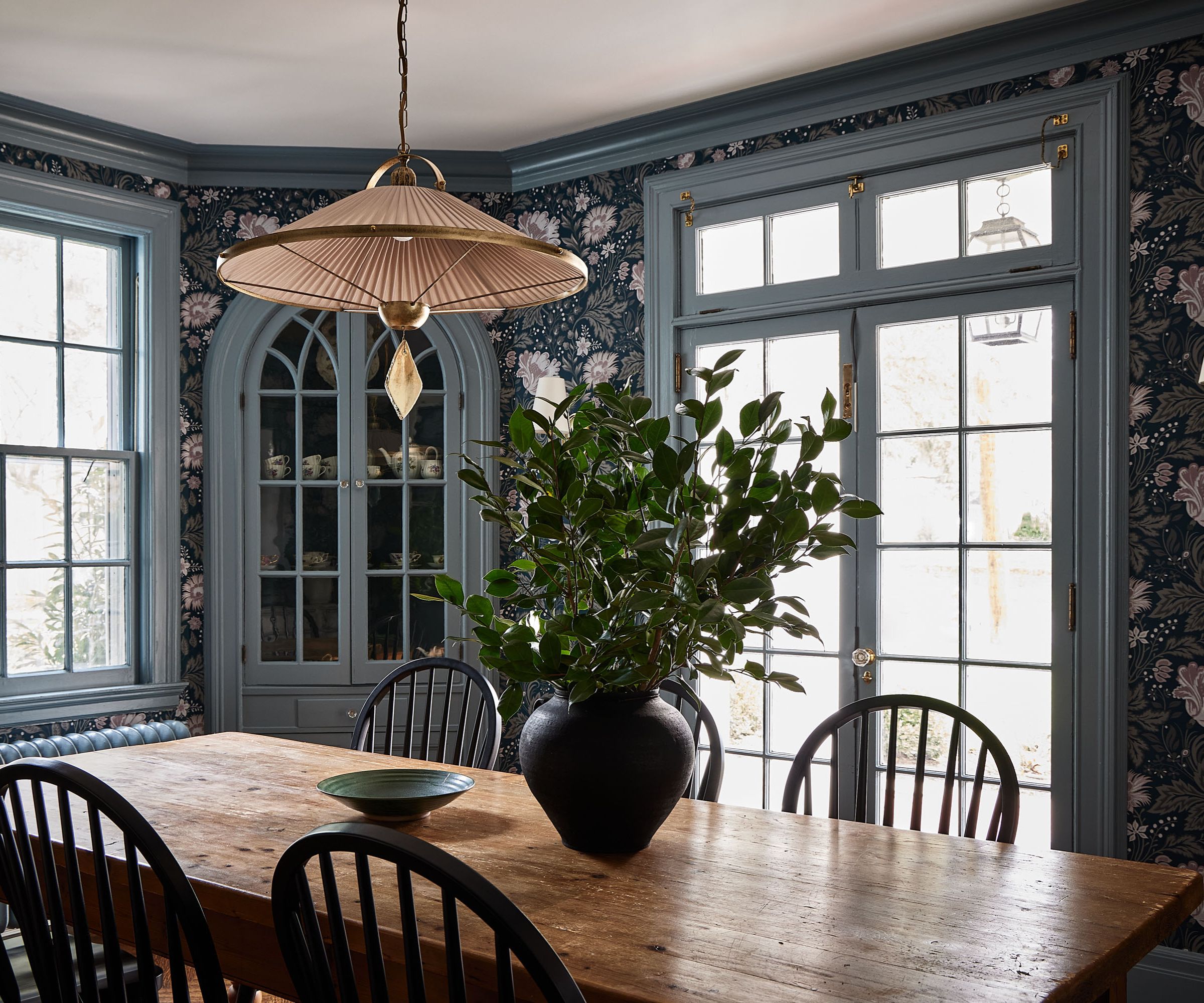 Dining room with floral wallpaper, blue painted door frame and window, long wood dining table and curved wood dining chairs and low gold pendant light