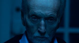 Saw X lead actor Tobin Bell looking at the camera at light up with blue lighting