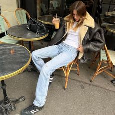 Camille wears a black coat with fur trim, white t-shirt, jeans, and adidas sambas.