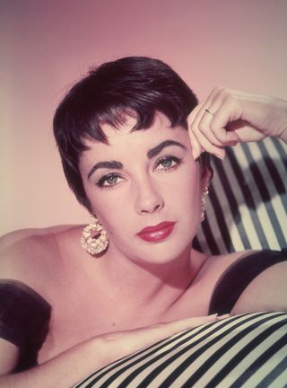 Promotional headshot portrait of British-born actor Elizabeth Taylor leaning on a striped sofa in a sleeveless gown and short hair