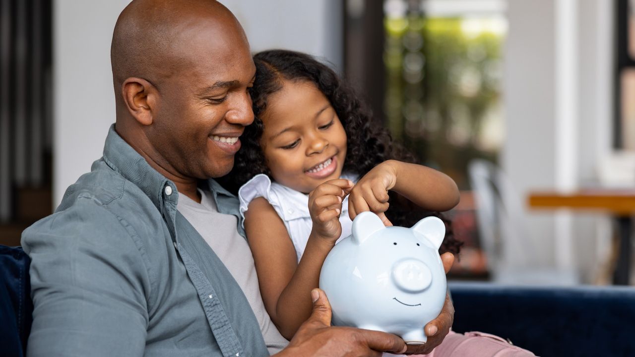 Daughter sitting on her father&#039;s lap, putting coins into a piggy bank as both of them smile