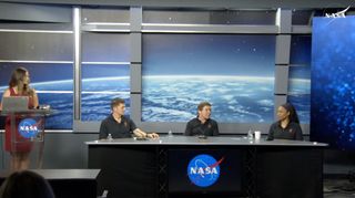 three people in dark blue polo shirts sit at a table during a press conference