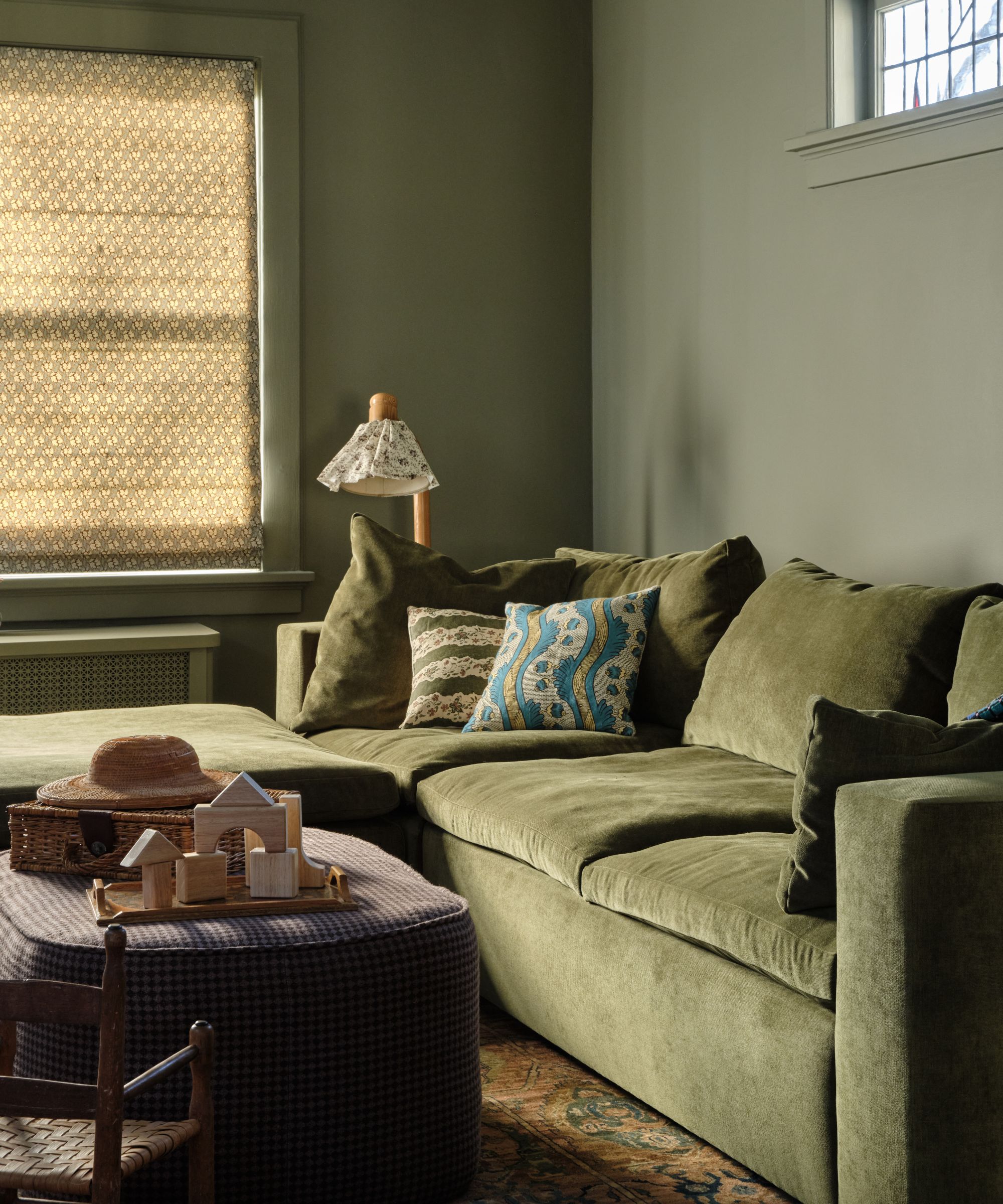 a moody green color drenched family room with a green couch, floral sofa pillows, an ottoman and a small vintage floor lamp