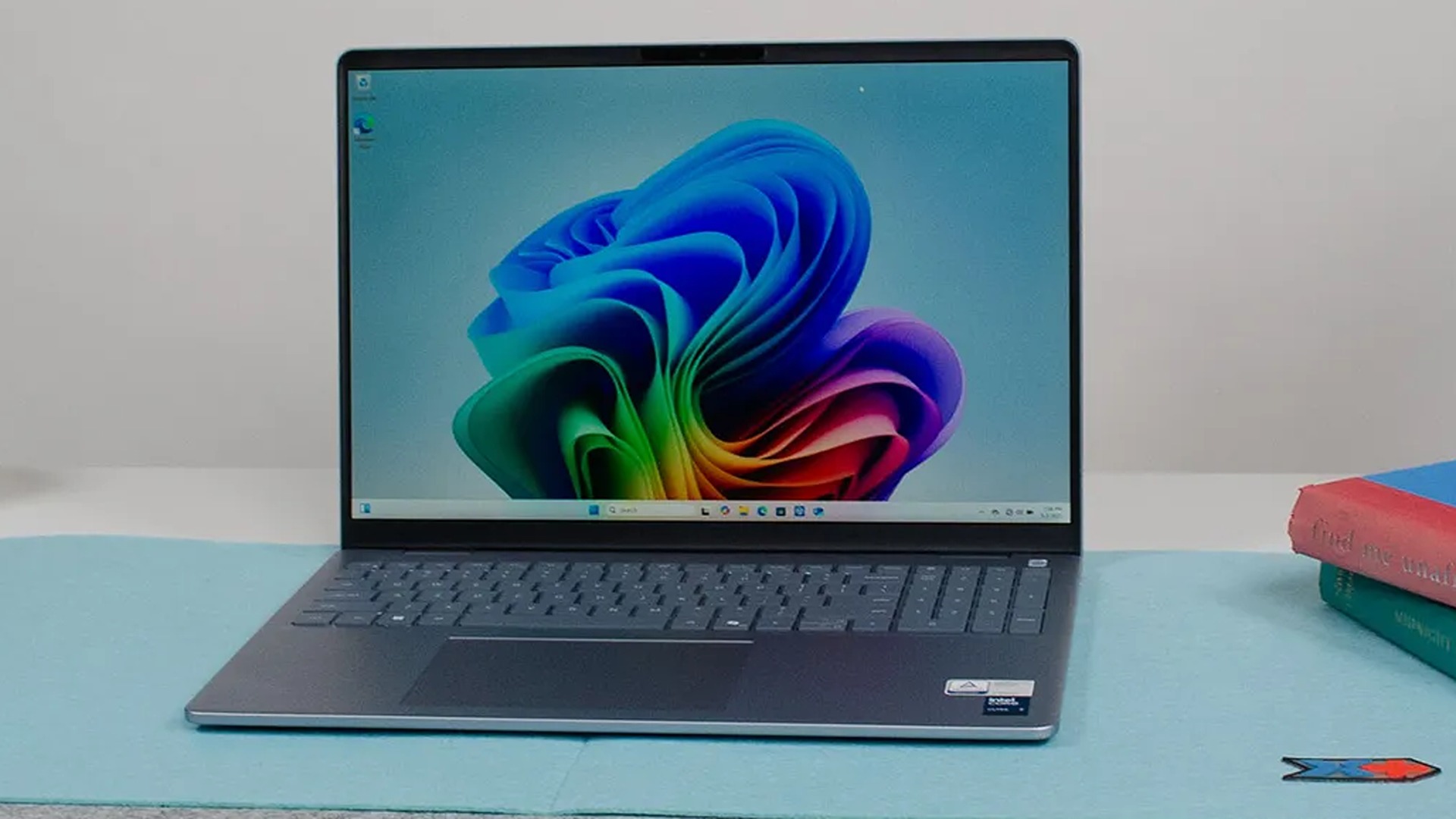 A sleek laptop with a vibrant blue and green abstract wallpaper on the screen, placed on a light blue surface next to colorful books.