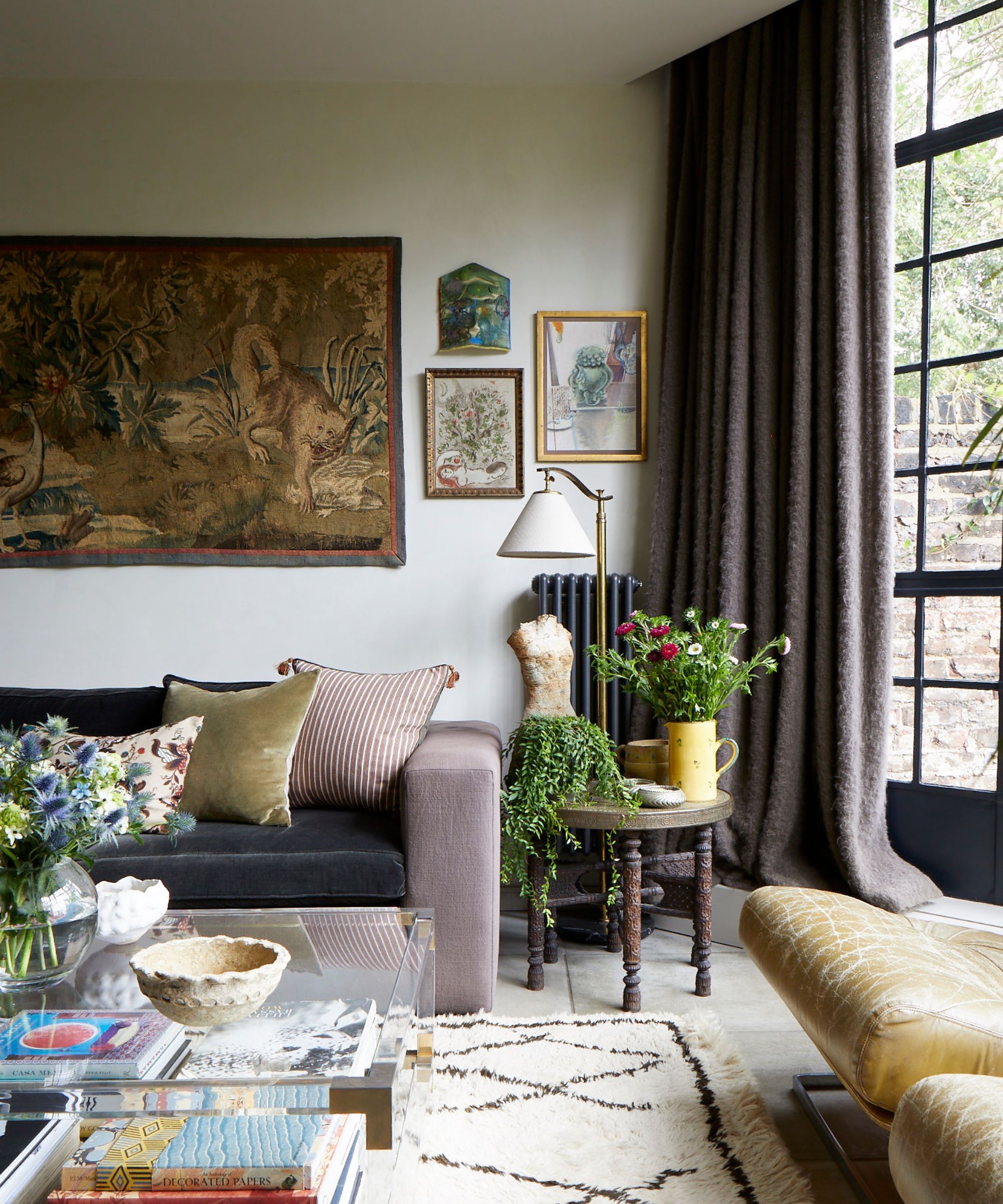 An eclectic, cozy living room featuring a velvet sofa with striped pillows, a large animal tapestry on the wall, and a glass coffee table layered with books and flowers