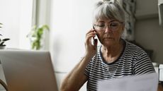 An older woman has a serious expression while she talks on the phone and looks at paperwork.