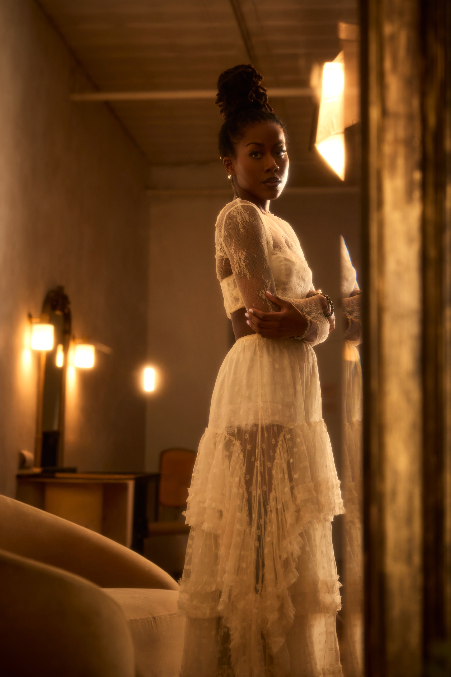 Author Tomi Adeyemi wears a white semi-sheer dress while standing in a room with an armchair and lit wall sconces.