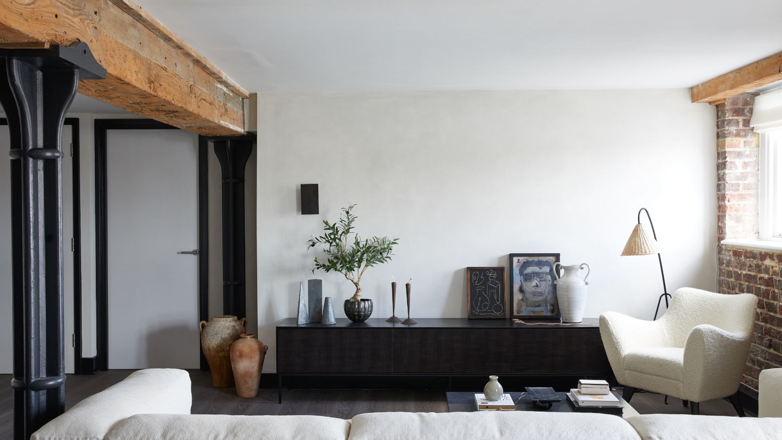 Industrial loft style living room with exposed beams and brick wall, plaster walls, console table with artwork and potted plant, boucle chair, white sofa and floor lamp