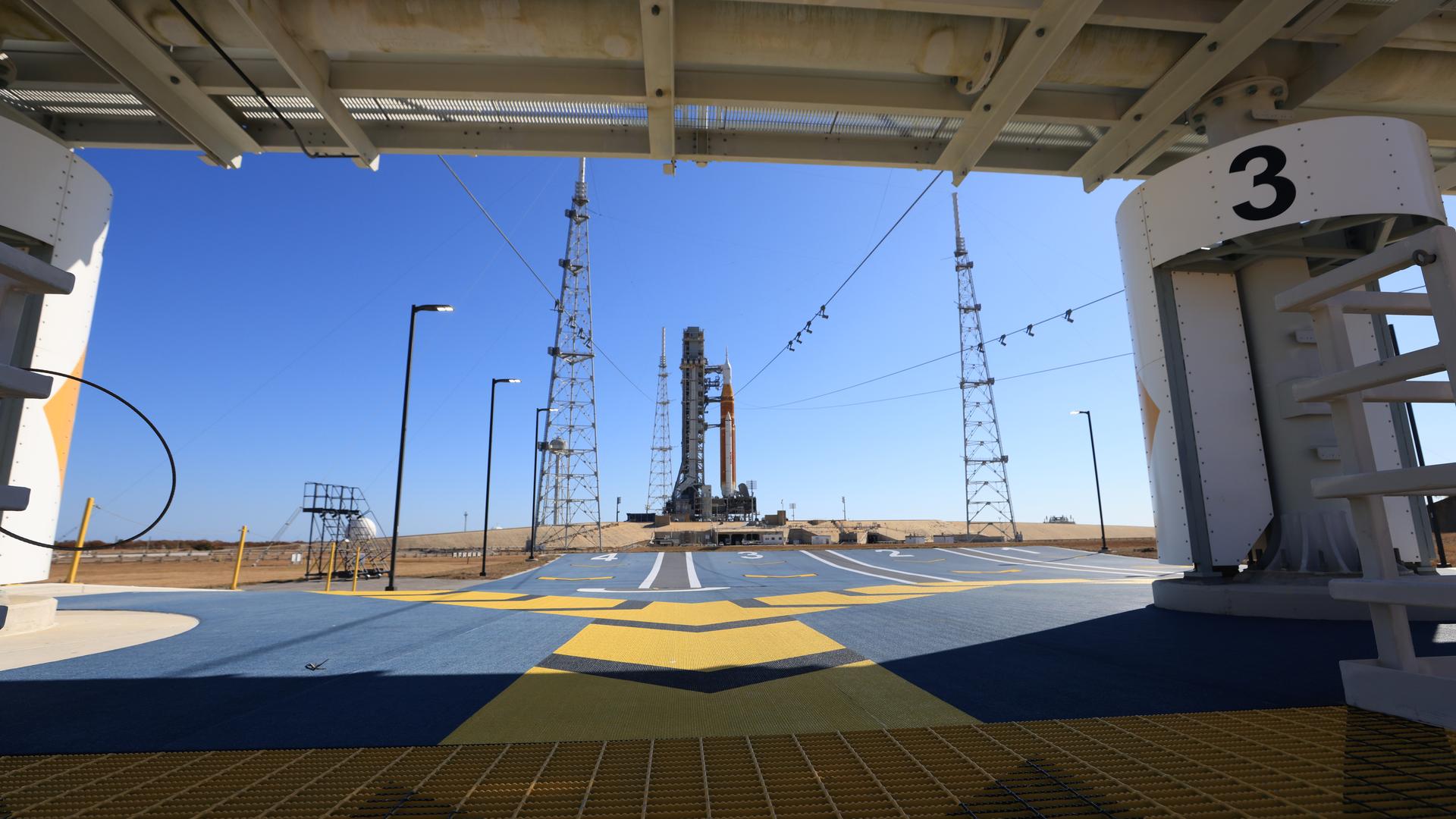 A white and orange NASA moon rocket on its launch pad as seen from an ermegency escape base conected by zip lines to the rocket gantry