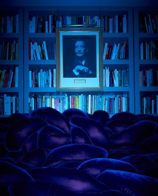 Portrait of Salvador Dalí hanging above navy sofa with book shelf behind