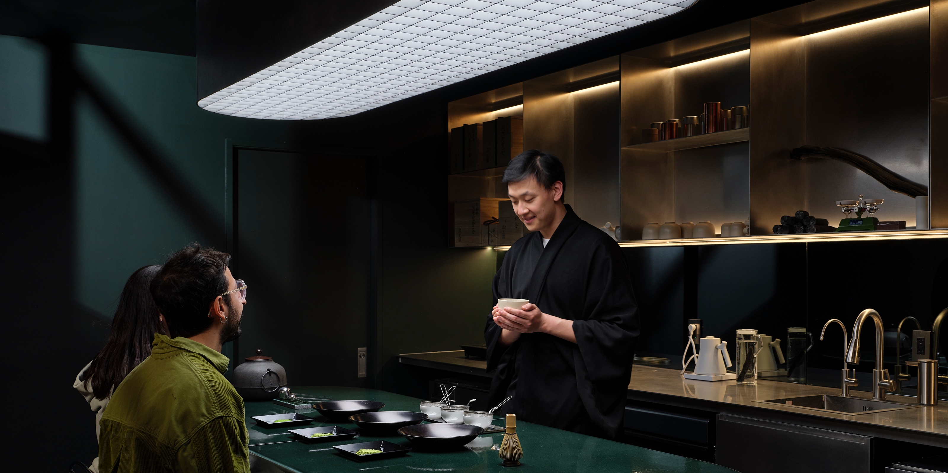 A cinematic matcha cafe features a sculptural, floating pale white light with a grid-like motif, a dark green countertop, chrome finishes, and wood accents, as a barista and two people enjoy a matcha ritual.