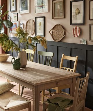 Dining room with pale pink painted walls and black painted paneling, feature wall of framed pictures and decorative objects, light wooden rectangular dining table with wooden dining chairs in different finishes, vases and jugs on dining table