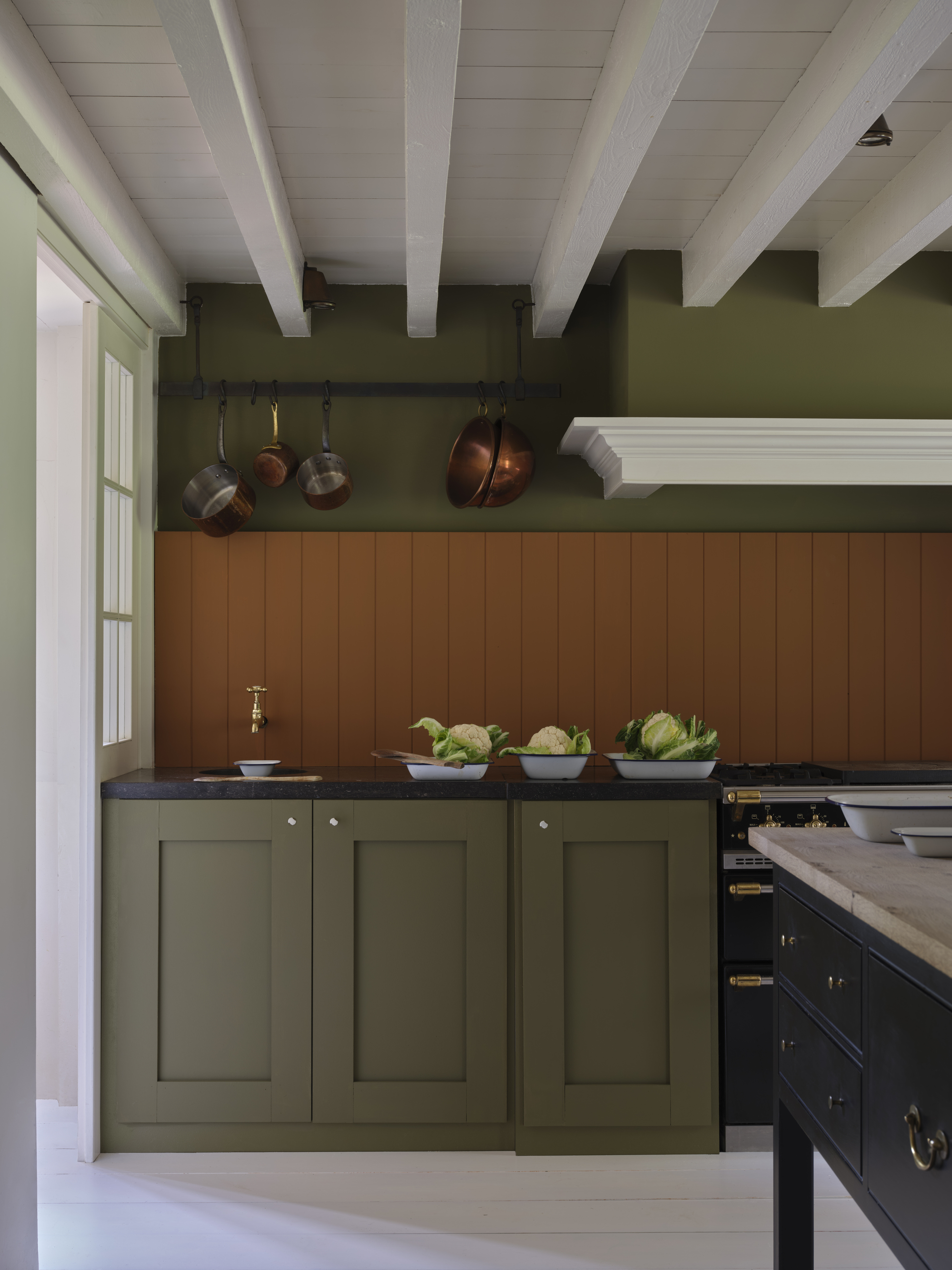 Image of a modern kitchen with white ceilings, olive green walls and cabinetry, and a burnt orange wood-paneled backsplash. There are pots on a hanging rail and a stand-alone kitchen island also in the room.