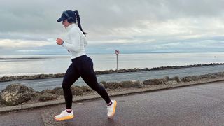 A girl in a white top and black leggings, cap and headphones running along road by sea