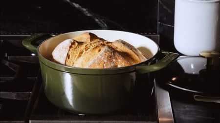 A deep green Made In Dutch Oven on a gas stove with a freshly baked loaf of bread inside. The lid sits beside it on a grey counter