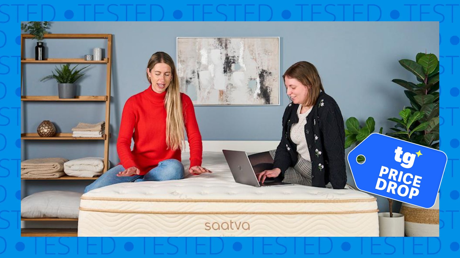 Two mattress testers talk while sitting on top of a cream and gold Saatva mattress, which is a placed on a white bed frame within a neutrally decorated bedroom. Inset, a sale tag reads 'TG price drop'