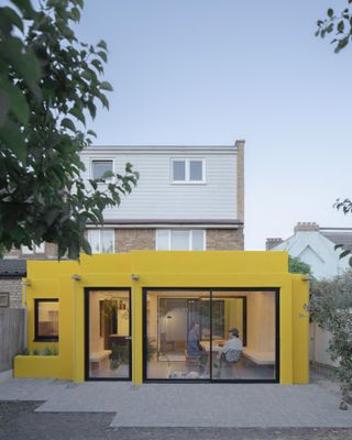 Behind another building is a yellow studio with patio windows/doors and two people inside.