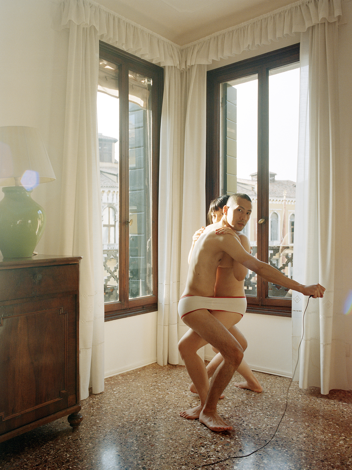 In a sunlit room with tall windows overlooking Venetian buildings, a man in white briefs crouches in a wide stance while a woman presses close behind him, as he holds out a shutter release cable.