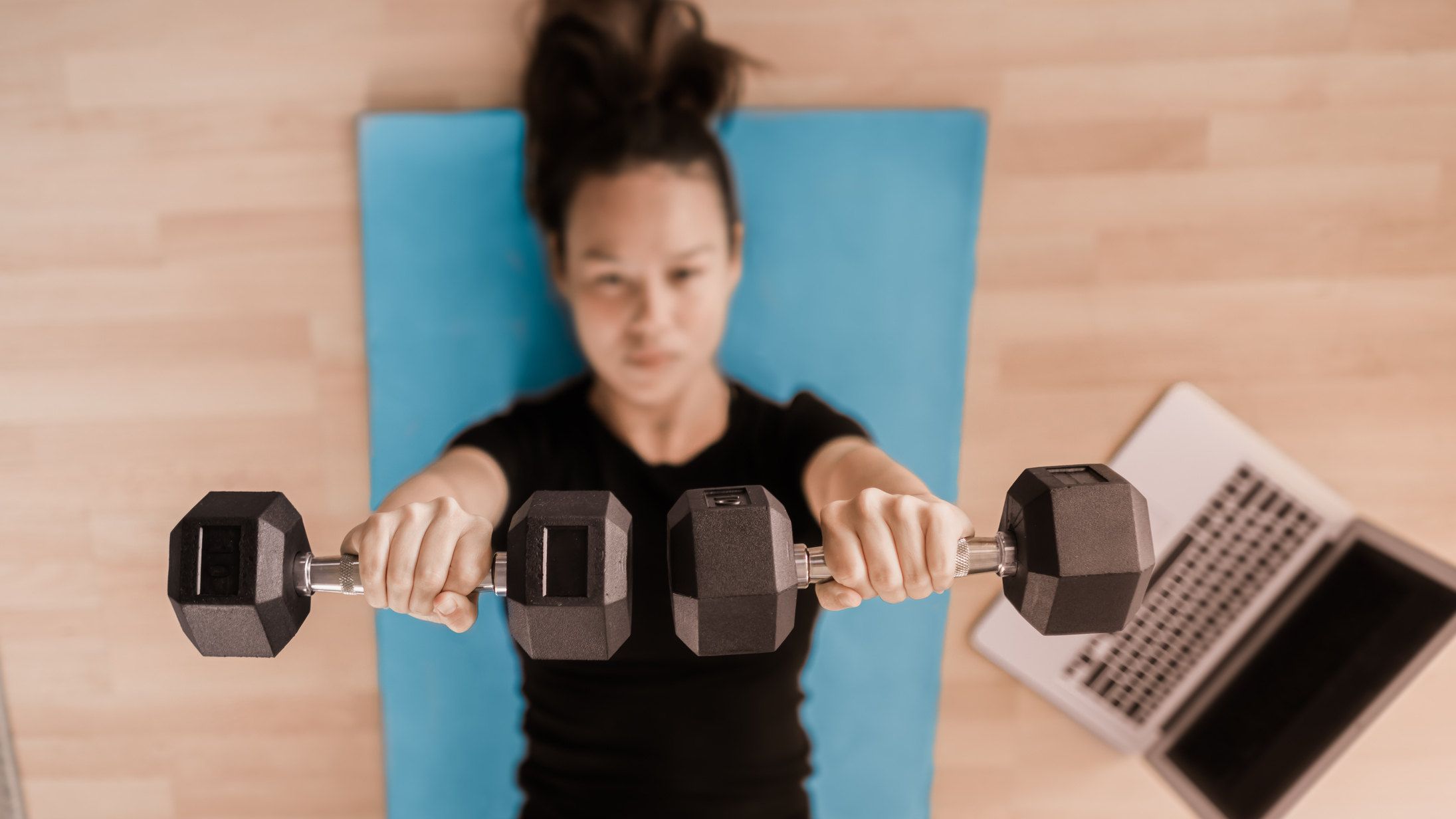 Woman lying on her back on an exercise mat holding dumbbells above her with straight arms. A laptop is on the floor next to her
