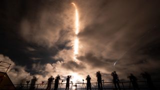 a white rocket lifts off at night above a plume of fire and smoke