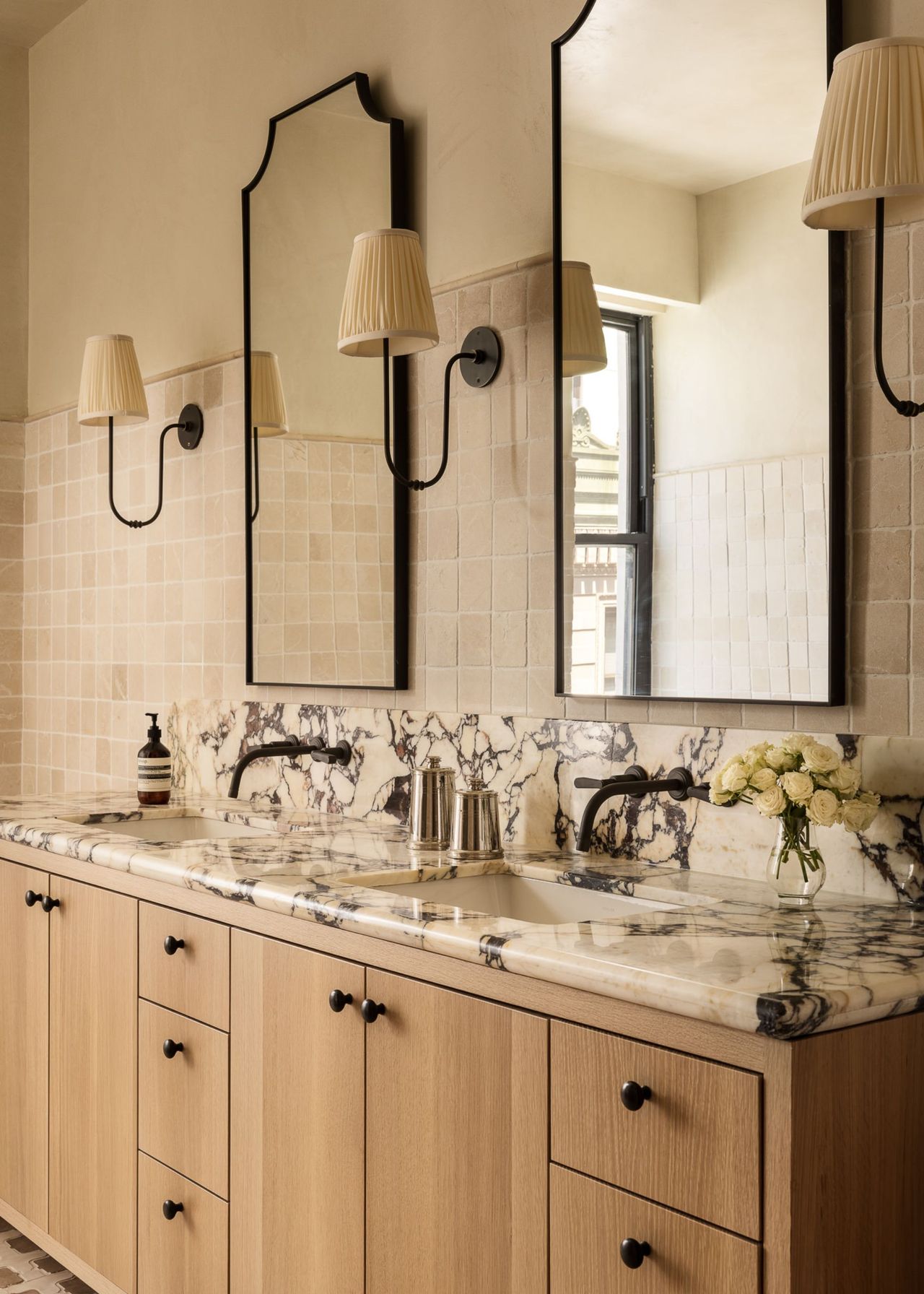 A modern bathroom with a double vanity with wooden cabinetry and marble counters, black wall-hung taps and two mirrors with three wall lights on the beige tiles