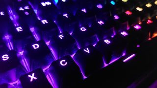 A close up of the Corsair K70 Pro TKL keyboard, showing the RGB lighting working in a darkened room. Many of the keys are lit up purple, though some keys can be seen in the distance in teal, red, and yellow.