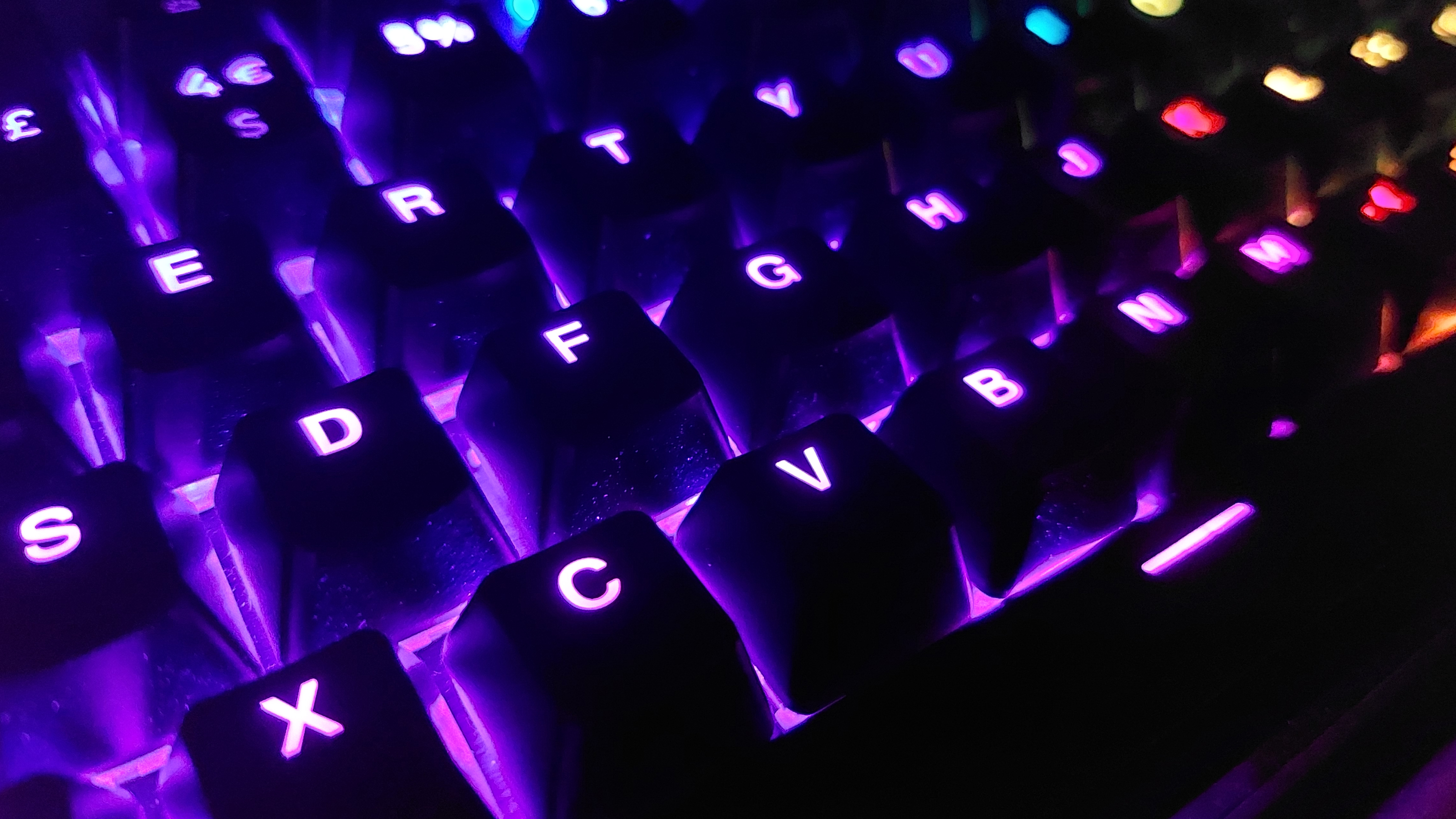 A close up of the Corsair K70 Pro TKL keyboard, showing the RGB lighting working in a darkened room. Many of the keys are lit up purple, though some keys can be seen in the distance in teal, red, and yellow.