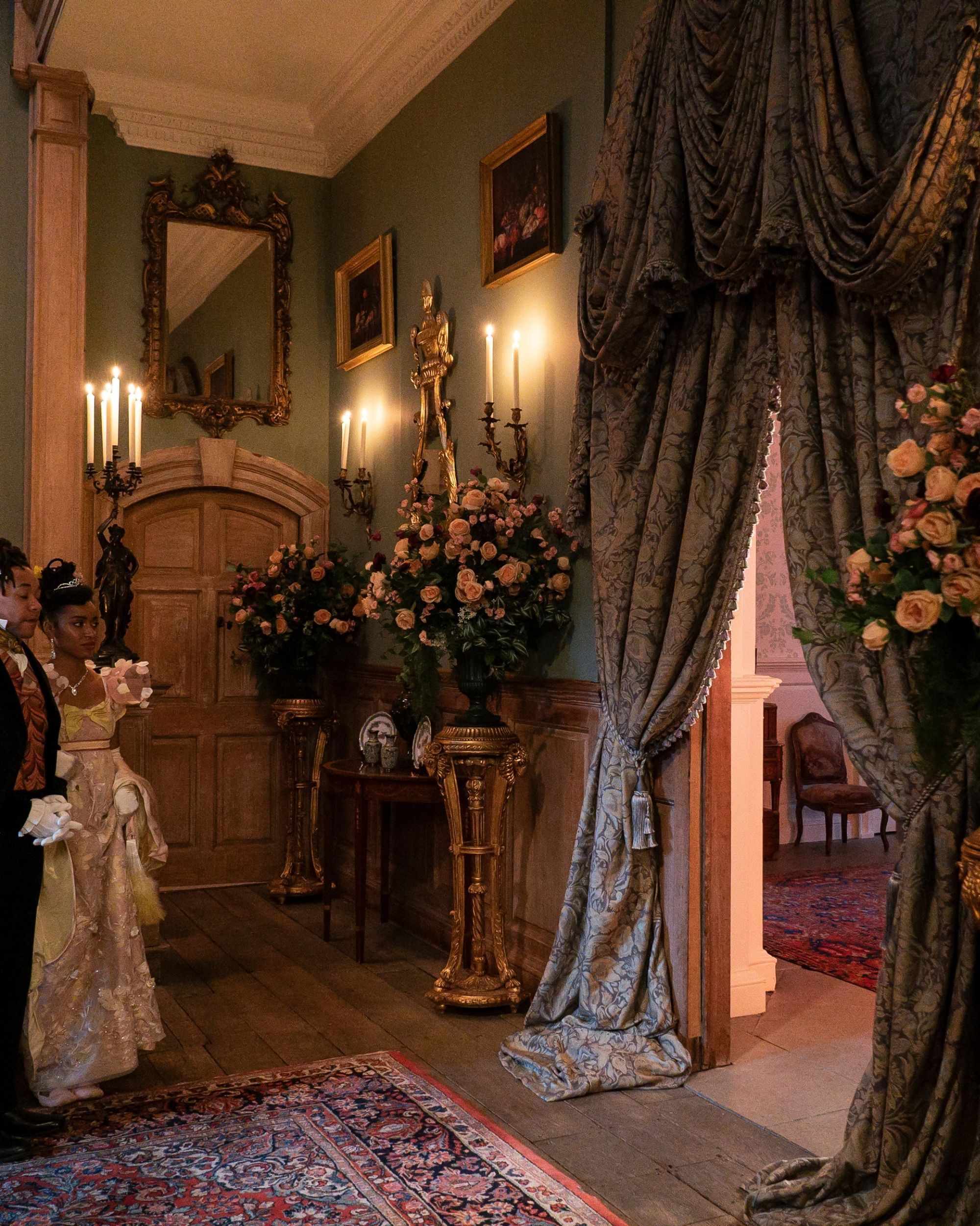 Production still from Netflix's Bridgerton series of a grand hallway/entryway that is blue with large, heavy, blue drapery framing the doorway. There are candle sconces, vases of flowers, and picture frames in the space as well.