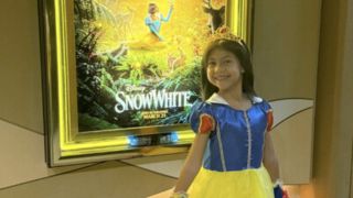 Maia Kealoha wearing a Snow White Disney Princess costume with a matching tiara smiling in front of the Snow White movie poster