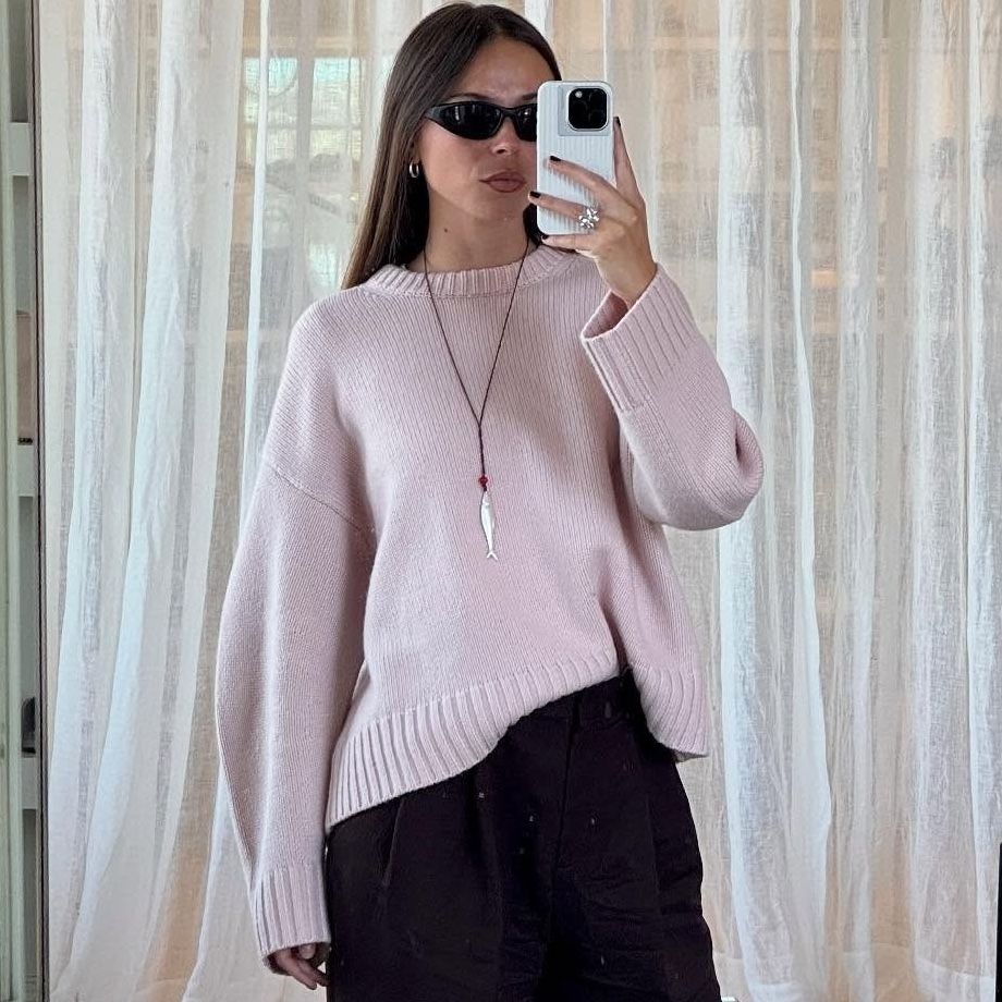 Scandinavian fashion creative Johanna Lager poses for a mirror selfie wearing black sunglasses, a chunky pink crewneck sweater, a red cord necklace with a silver fish pendant, and black pants
