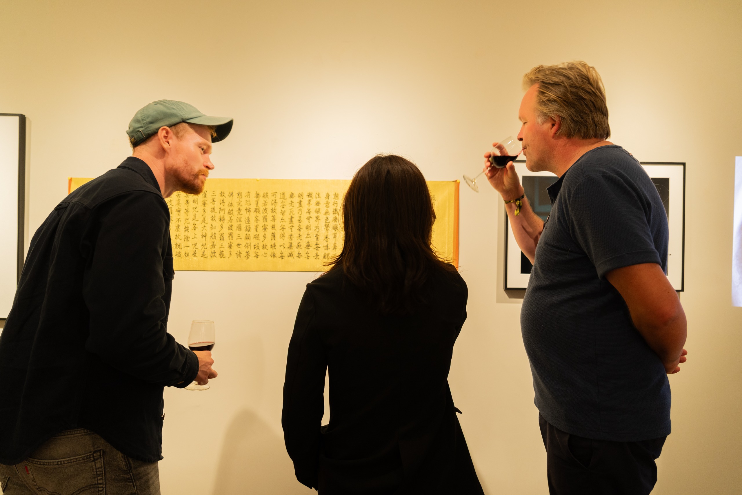 Three visitors, two holding glasses of red wine, lean in to study a long yellow panel of Chinese calligraphy on a gallery wall.