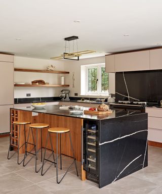 A kitchen island with a black marble counter with a bowl of fruit and chopping board on it, a wooden base, three circular wooden bar stools underneath, and pale pink cabinets around it